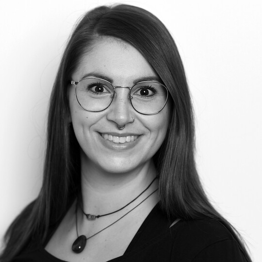 Porträt von Nathalie Benne. Sie trägt eine Brille, hat langes braunes Haar und lächelt in die Kamera. Im Hintergrund eine helle, neutrale Wand.